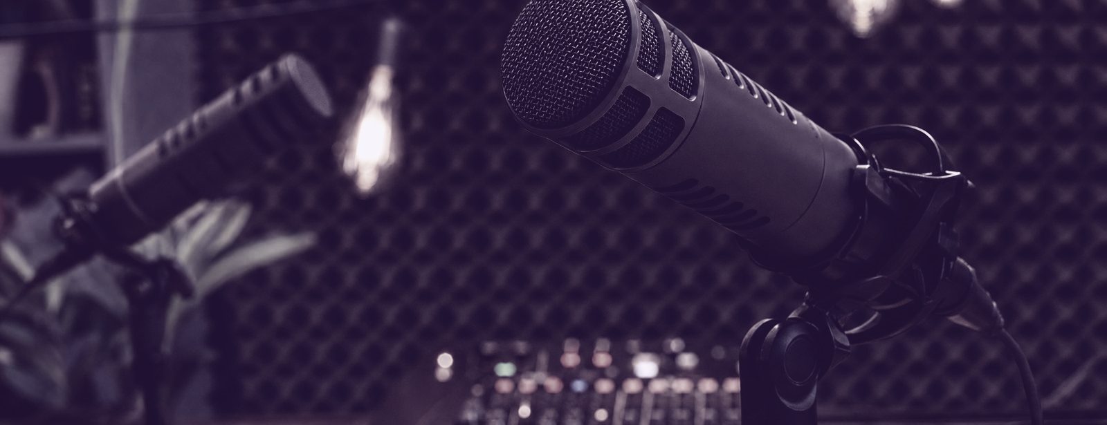 Two broadcast microphones on booms across a podcast desk, lit against acoustic foam